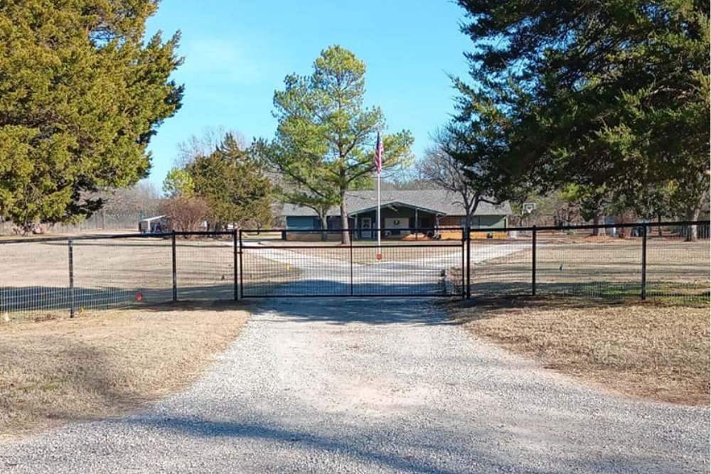 Exterior view of a home in the background with Amercian Flag, foreground has a solid well built heavy-duty gate and fence that surrounds the property. Driveway forks in the center.